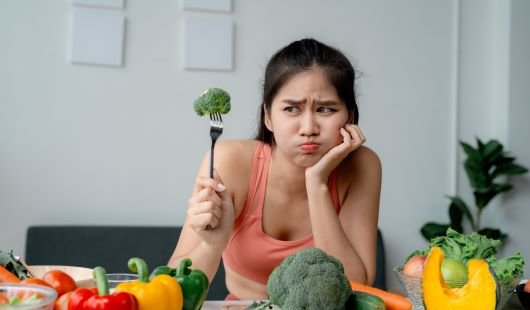 An unimpressed woman holding up a piece of broccoli on a fork.